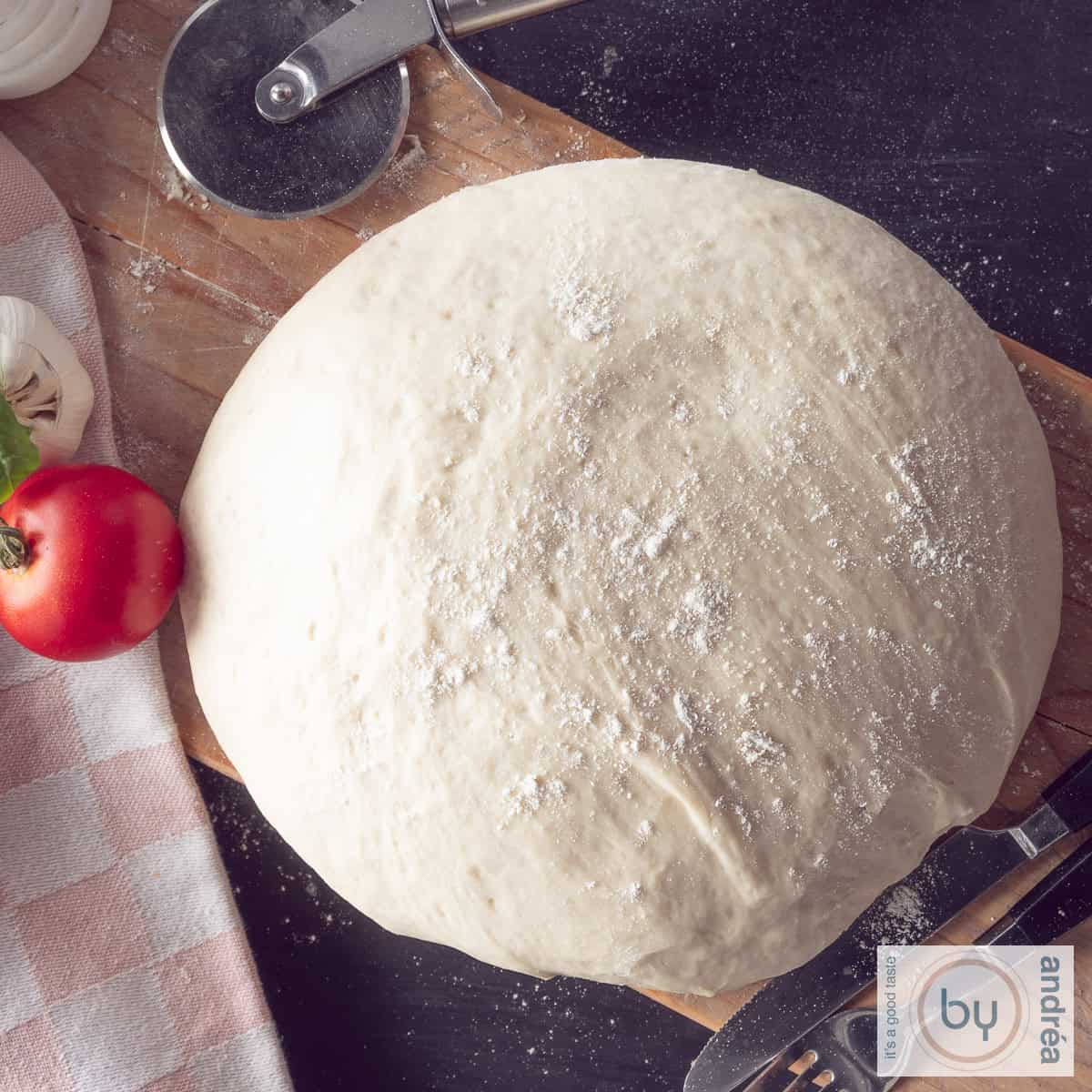 Een vierkante foto van bovenaf genomen van een bol pizzadeeg op een houten plank. Een tomaat en knoflook en een pizzasnijder liggen erbij.