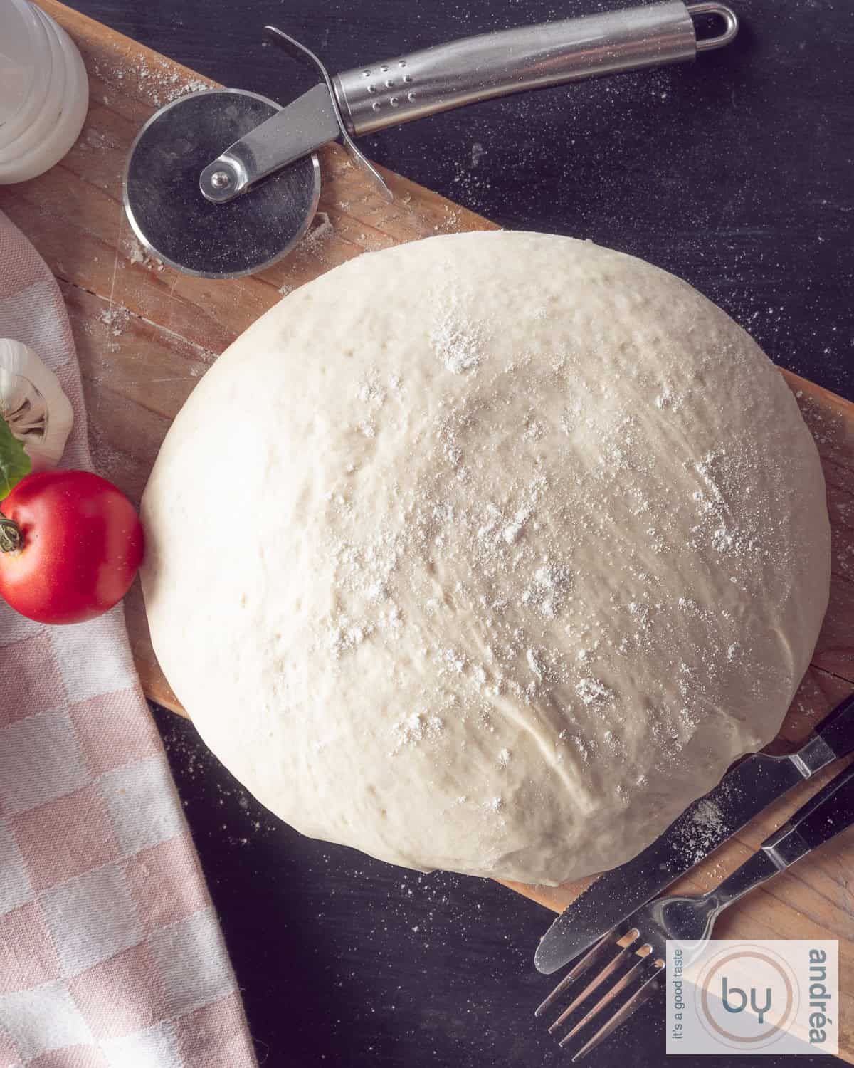 Een foto van bovenaf van een houten plank op een donkere ondergrond van een bol pizzadeeg. Bovenin ene pizzasnijder.