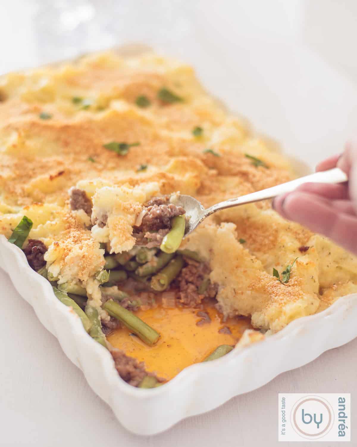 Een hoogte foto van een ovenschotel in een witte schaal met aardappelpuree, gehakt en boontjes. Een lepel pakt een schep eruit.