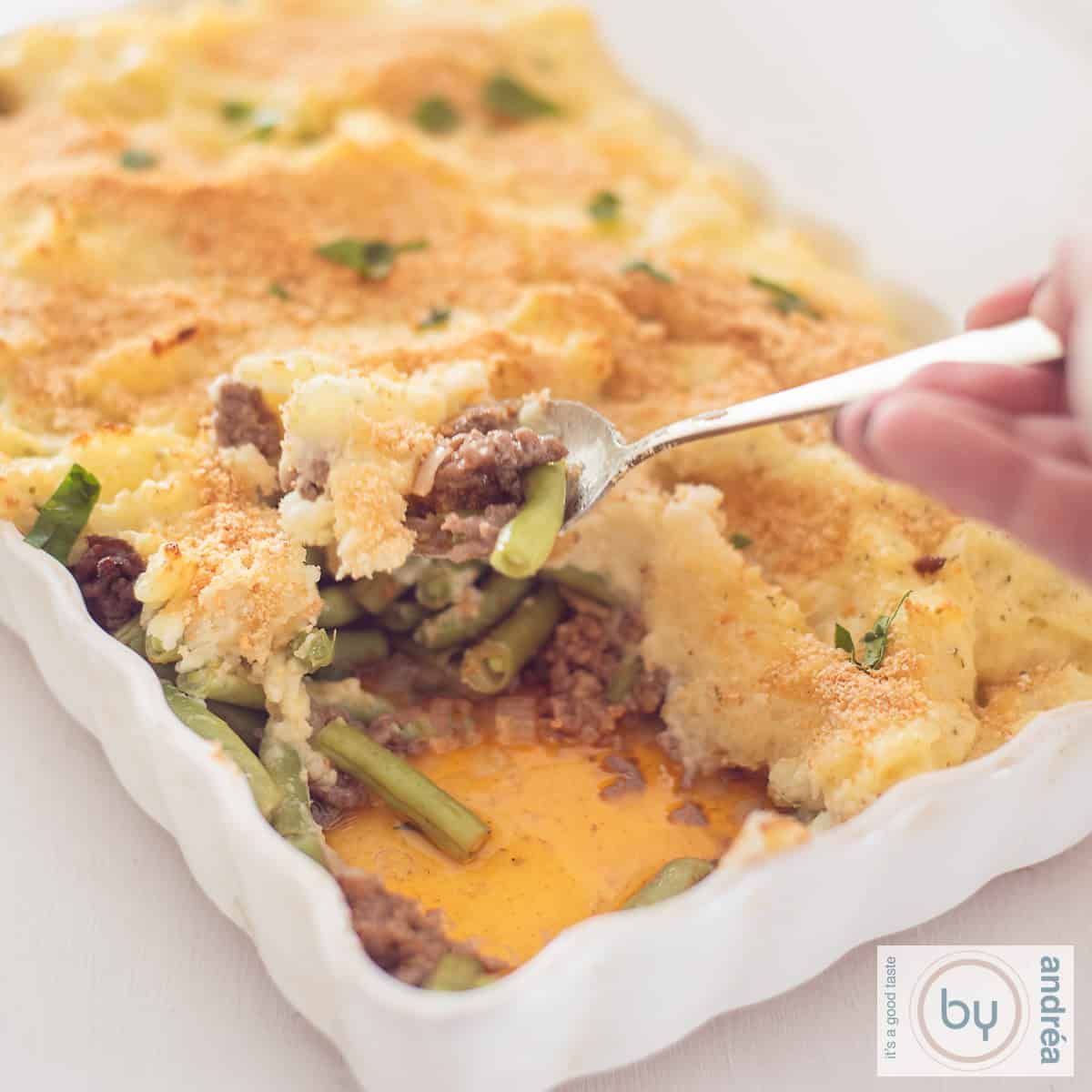 Een vierkante foto van een witte ovenschaal, waara een beetje uit is gehaald en de lagen boontjes, gehakt en aadappelpuree zichtbaar zijn.