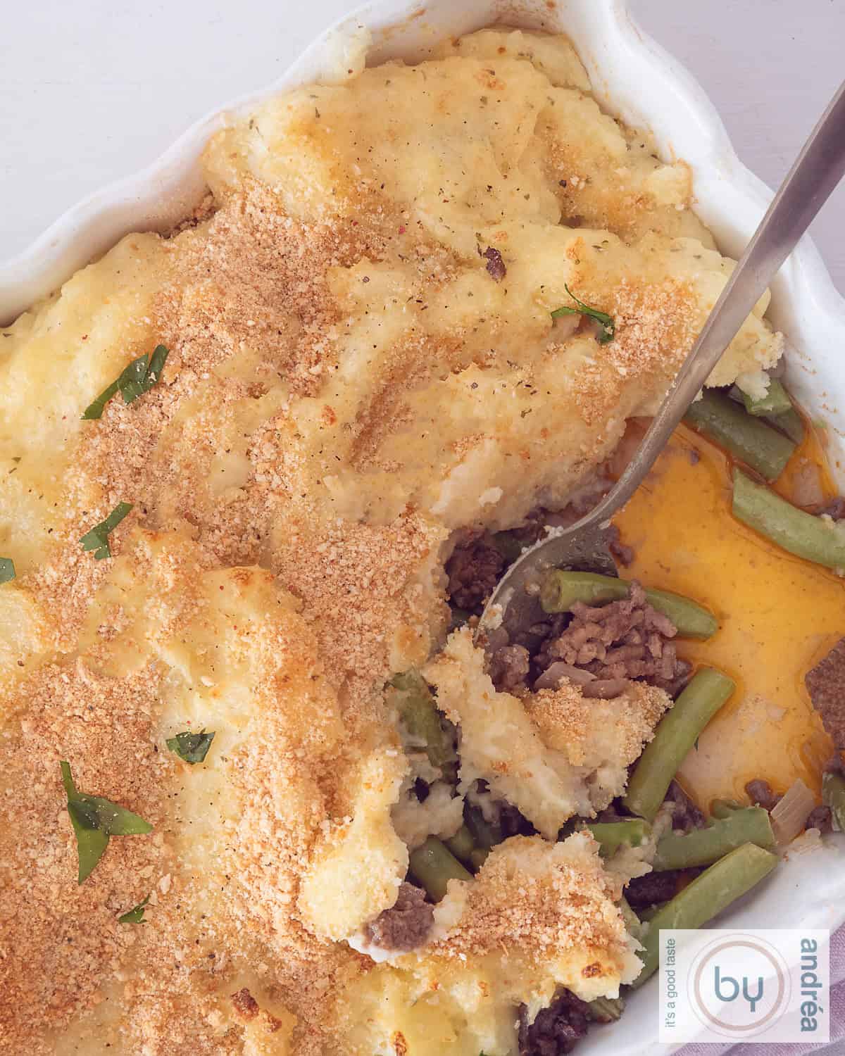 Een hoogtefoto bovenaf genomen van een ovenschotel met gehakt, boontjes en aardappelpuree. Een hap is eruit.