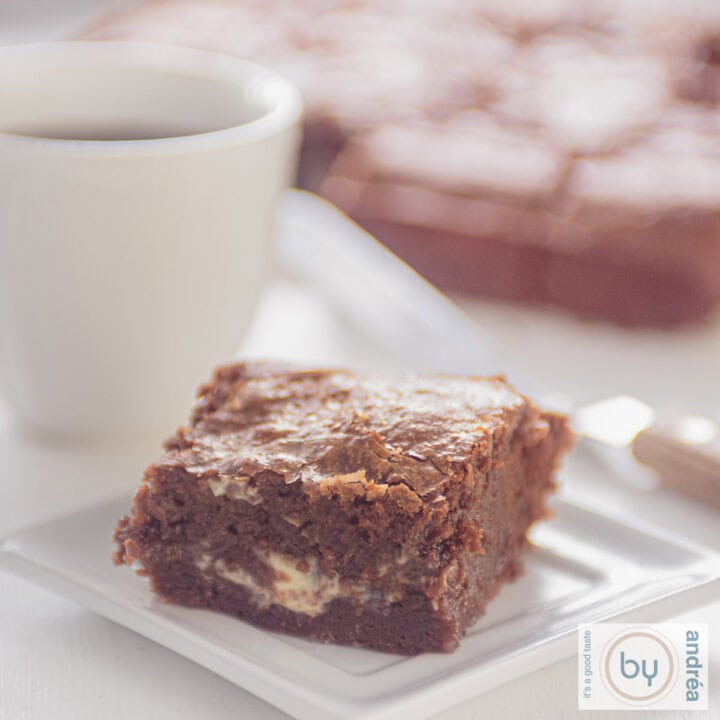 Een vierkante foto van een wit bord met een stukje bord met gesmolten witte chocola binnenin. Een kopje met koffie erachter samen met brownies.