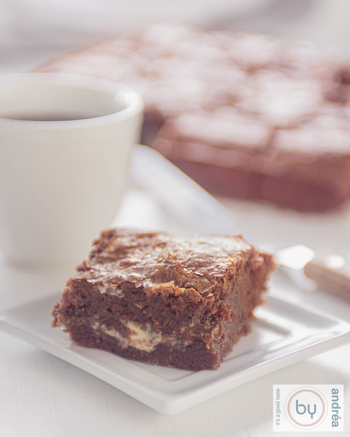 Een hoogtefoto van een wit bord met een vierkant stukje brownie gevuld met witte chocola. Een kopje koffie ernaast en meer brownies in de achtergrond.