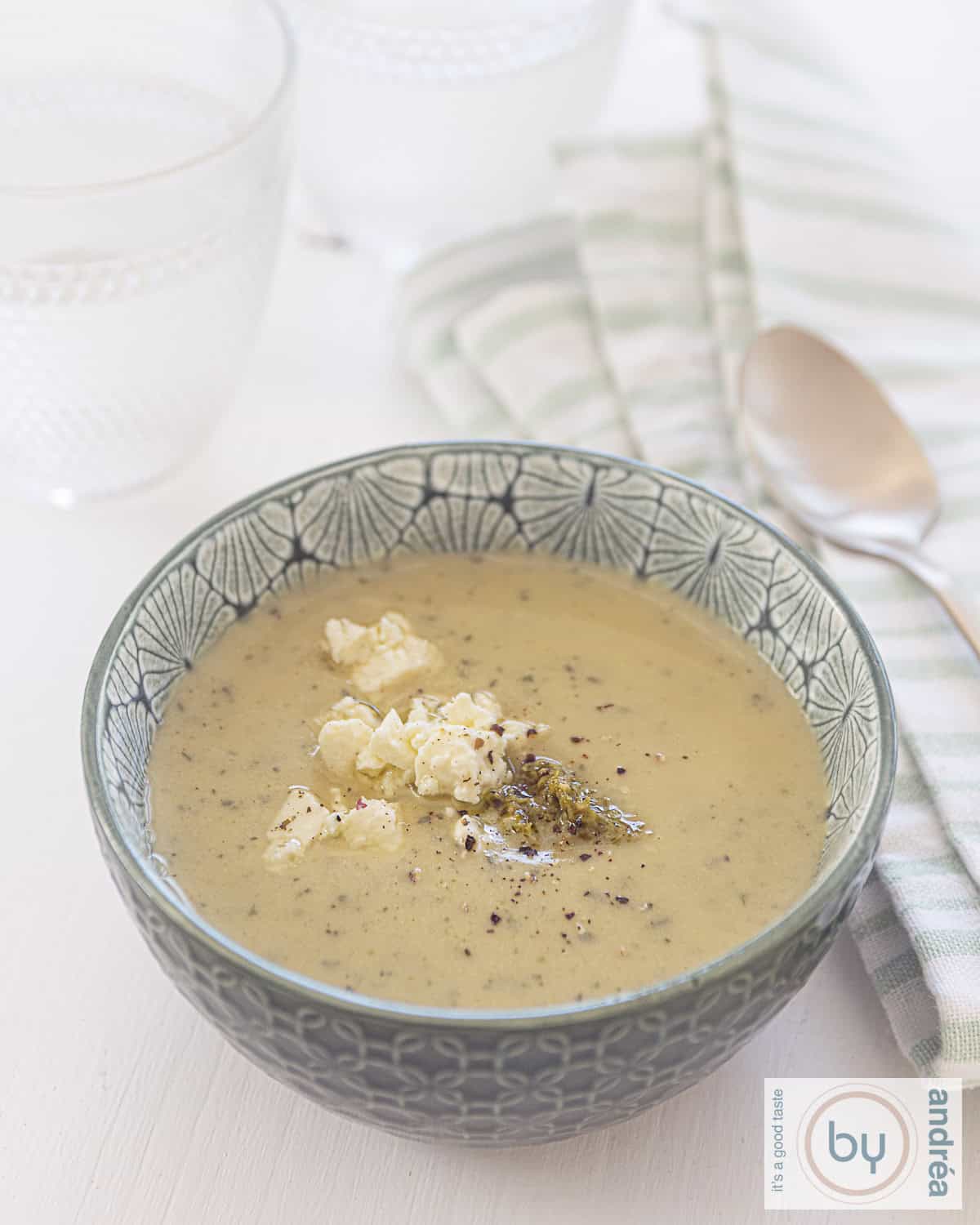 Een groene kom met courgetteosoep, feta en pesto op een witte ondergrond. Rechts een groen witte theedoek met daarop een gouden lepel