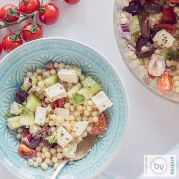 Een vierkante foto met twee schalen, een blauwe en een deel van een glazen schaal gevuld met salade van parelcouscous, komkommer, ui, feta en tomaatjes. Een trosje tomaten erboven.