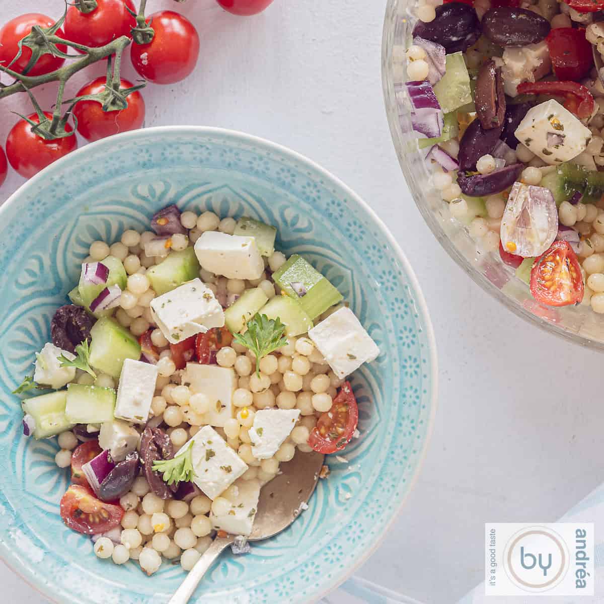 Een vierkante foto met twee schalen, een blauwe en een deel van een glazen schaal gevuld met salade van parelcouscous, komkommer, ui, feta en tomaatjes. Een trosje tomaten erboven.