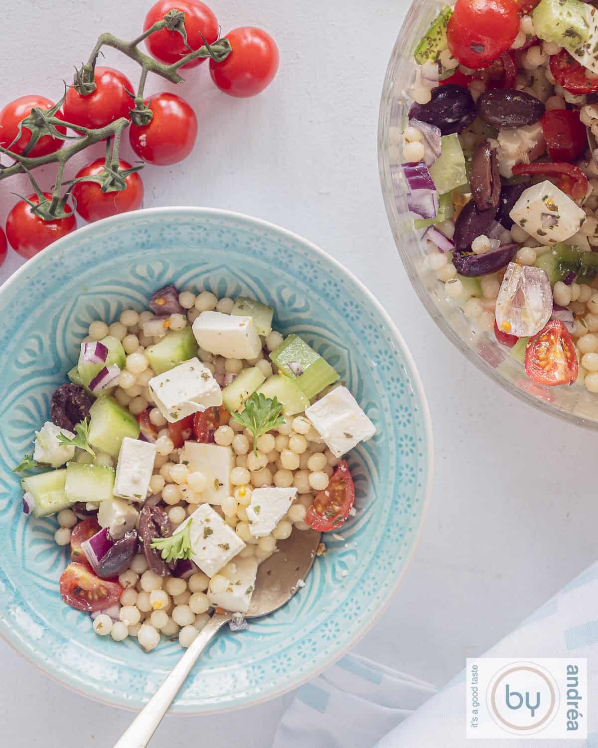 Een foto van bovenaf van een blauwe en een glazen schaal gevuld met parelcouscous salade met mediteraanse groenten en feta. Een trosje tomaten erboven