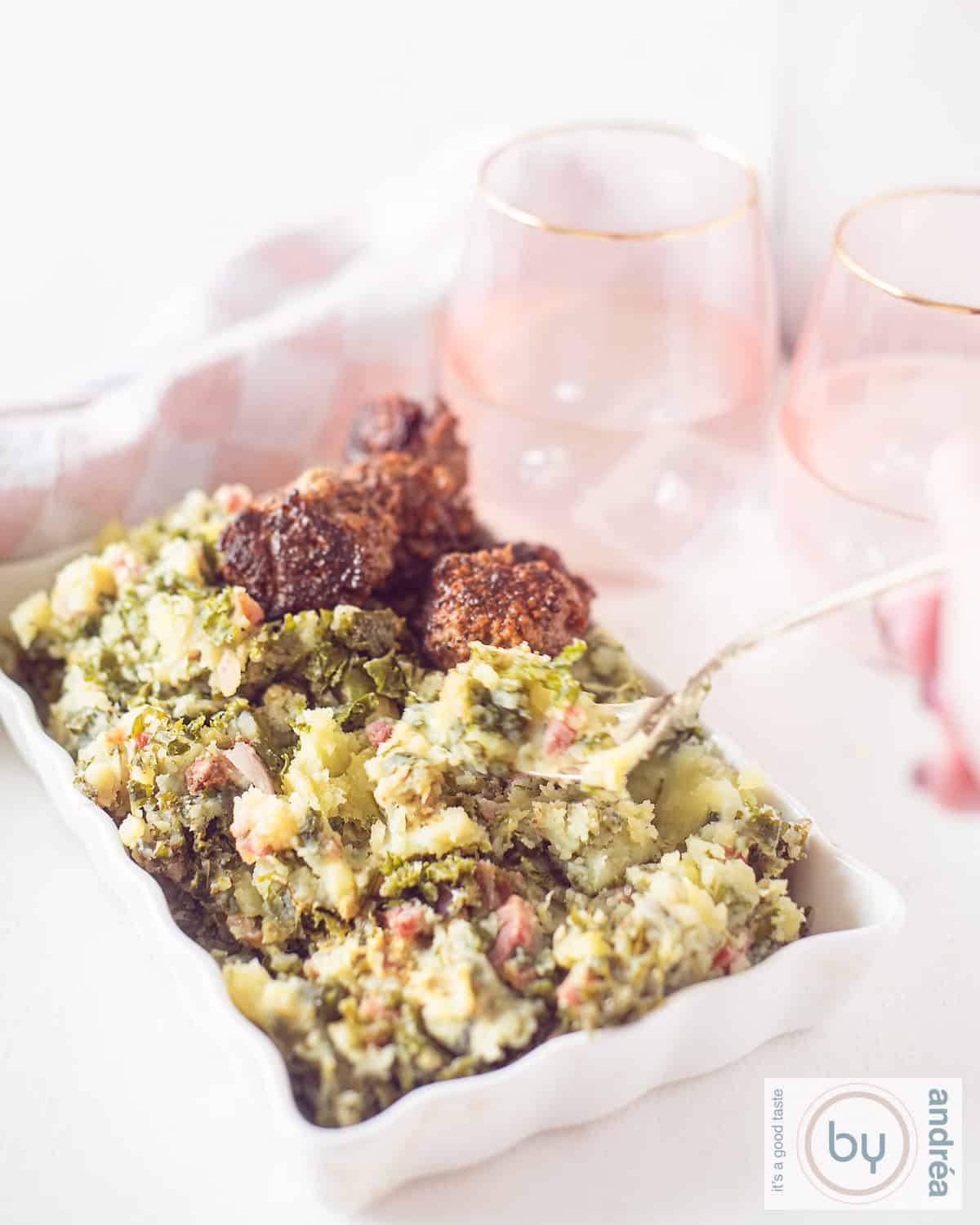 Een hoogtefoto van een witte ovenschaal met boerenkoolstamppot en gehaktballetjes. Een vork prikt eenhap eruit. Roze glazen in de achtergrond.