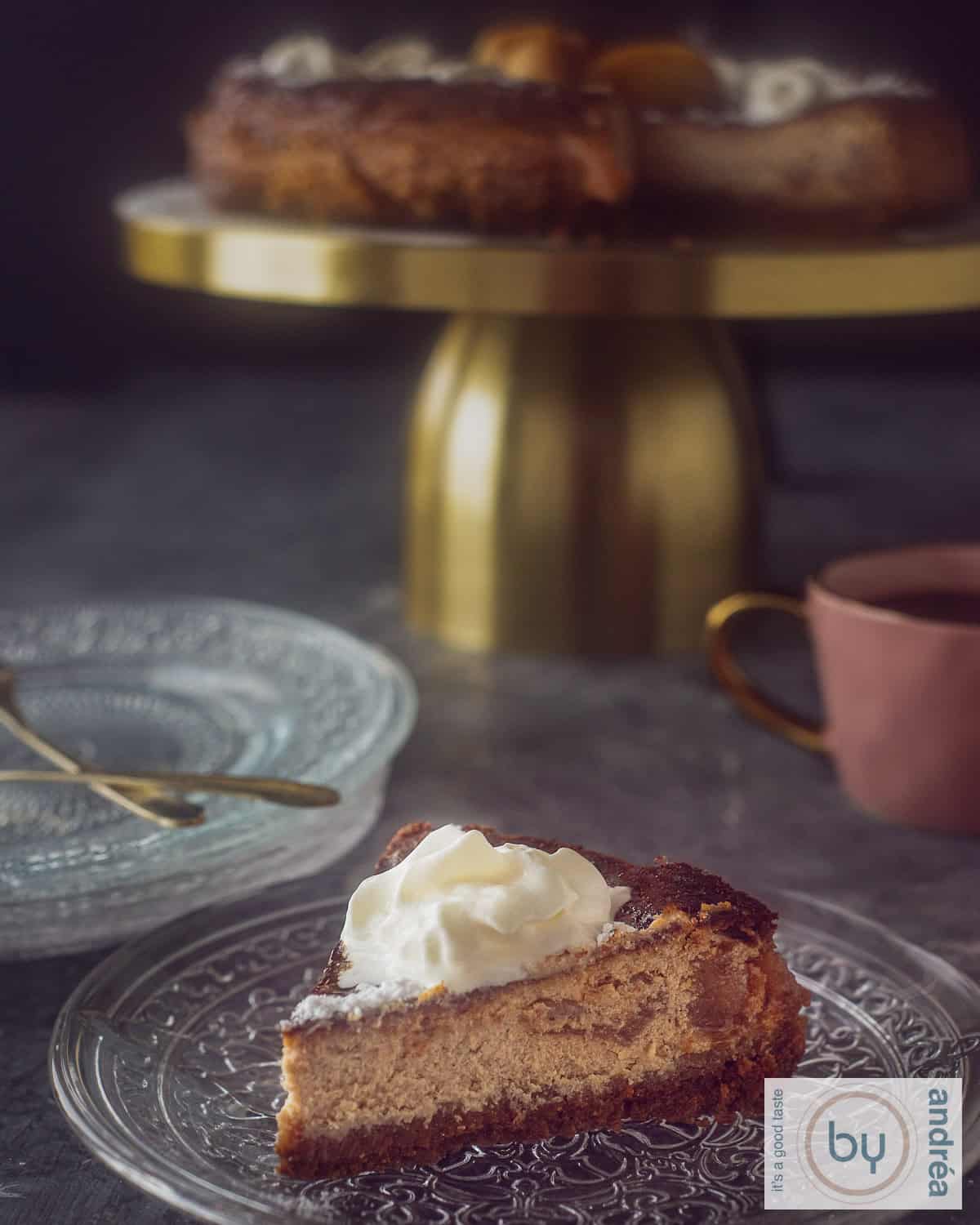Een stukje gekarameliseerde appel cheesecake op een glazen bordje met een toef slagroom. In de achtergrond een gouden taartstandaard met de rest van de cheesecake.