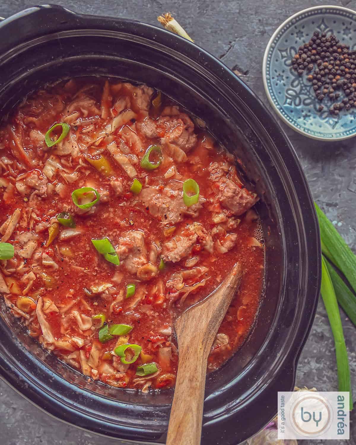 Een foto van bovenaf van een slowcooker met hamlappen, spitskool ine en heerlijke saus op een grijze ondergrond.