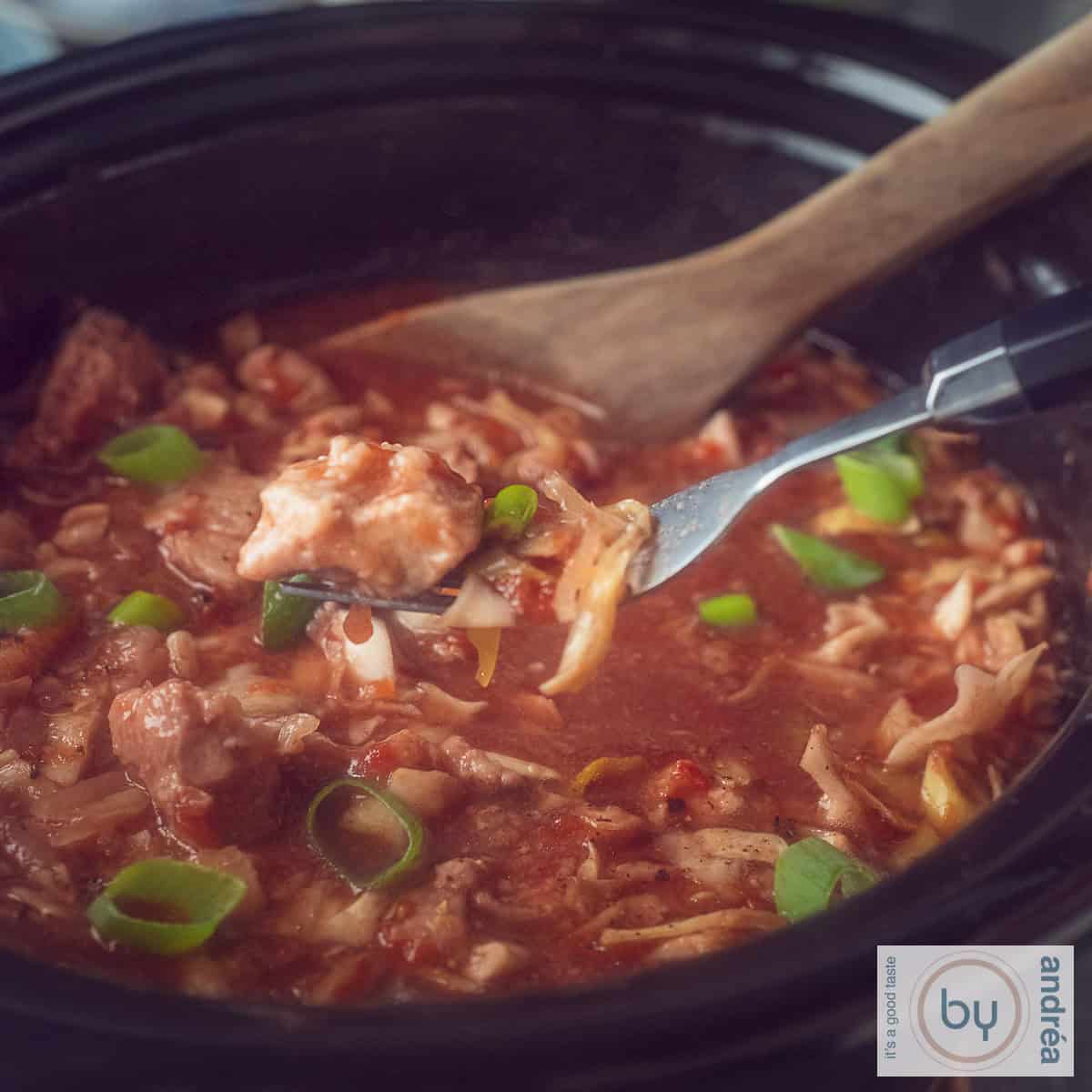Een vierkante foto met een deel van een slowcooker gevuld met spitskool, hamlappen en een rijke saus. Een vork pikt een stukje vlees uit de pot.