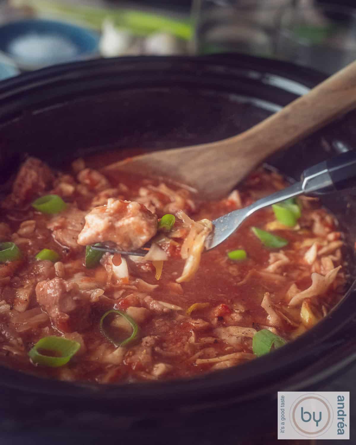 Een foto van een deel van een slowcooker gevud met hamlap, spitskool en een rijke saus. Een vork schept wat uit de slowcooker