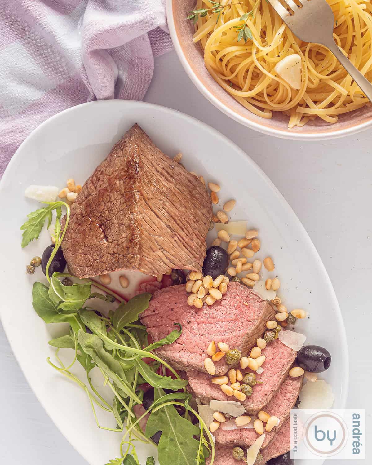 Een foto van bovenaf van Rosbief waar plakjes afgesneden zijn met pijnboompitjes, olijven, rucola en kaas. Een kom met linguine, knoflook en tijm rechtsbovenin