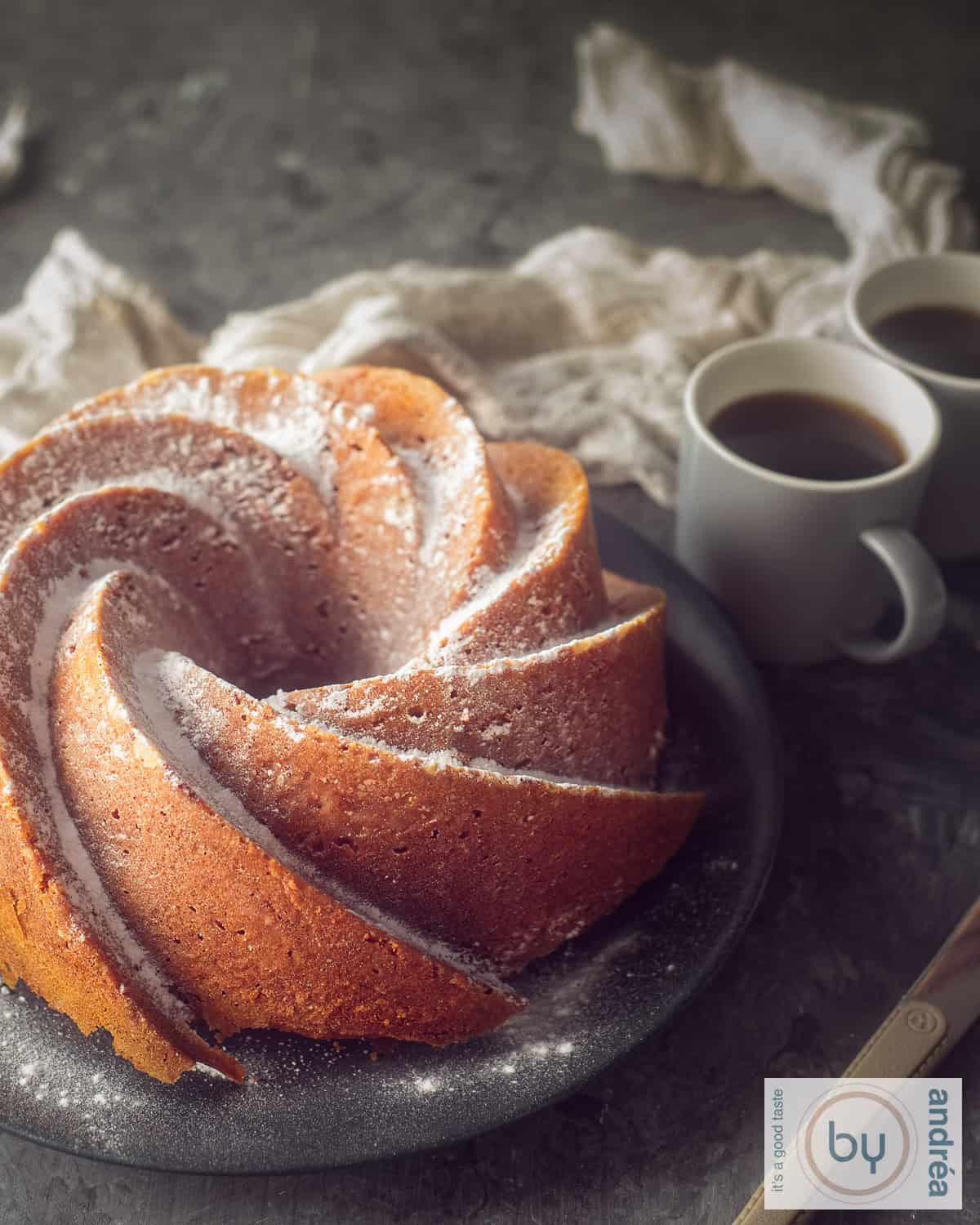 Een hoogtefoto een pompoen tulbandcake op een grijs bord. Twee kopjes koffie erachter een donkere ondergrond.