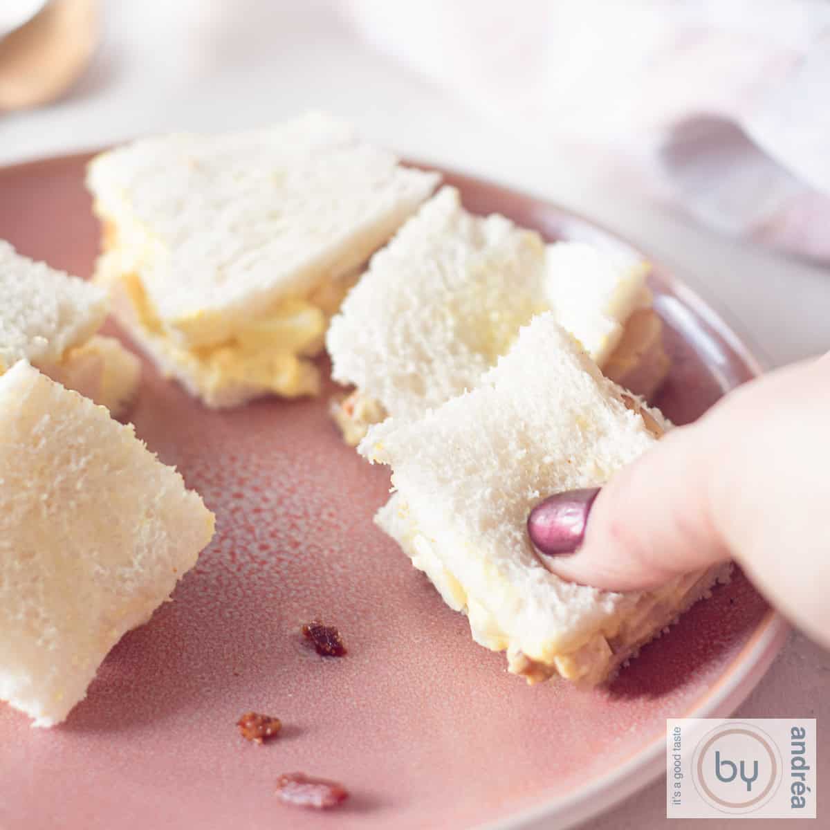 Een vierkante foto met een deel van een roze bord met daarop sandwiches met gerookte kip kerrie salade met spekjes en ei. Een hand pakt een sandwich.