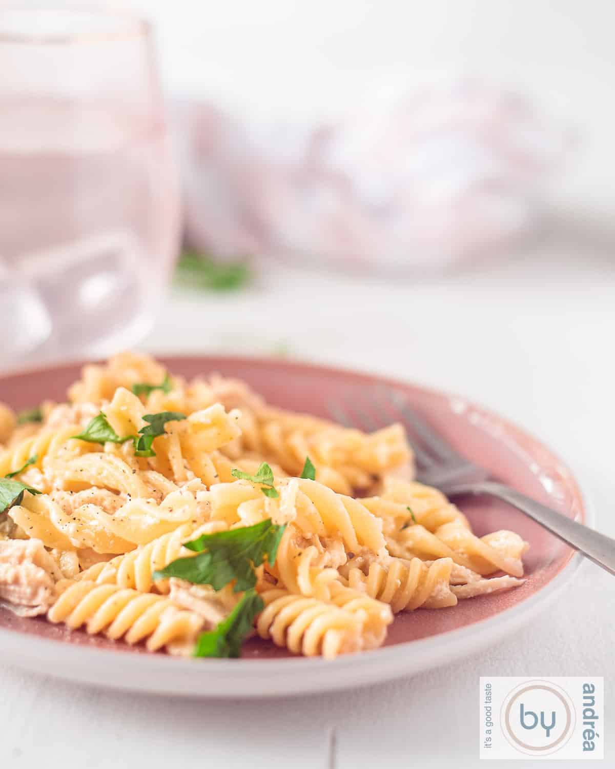 Een hoogtefoto met een roze bord met fusilli met kip, knoflook parmesaan saus, peterselie. In de achtergrond roze glazen met water