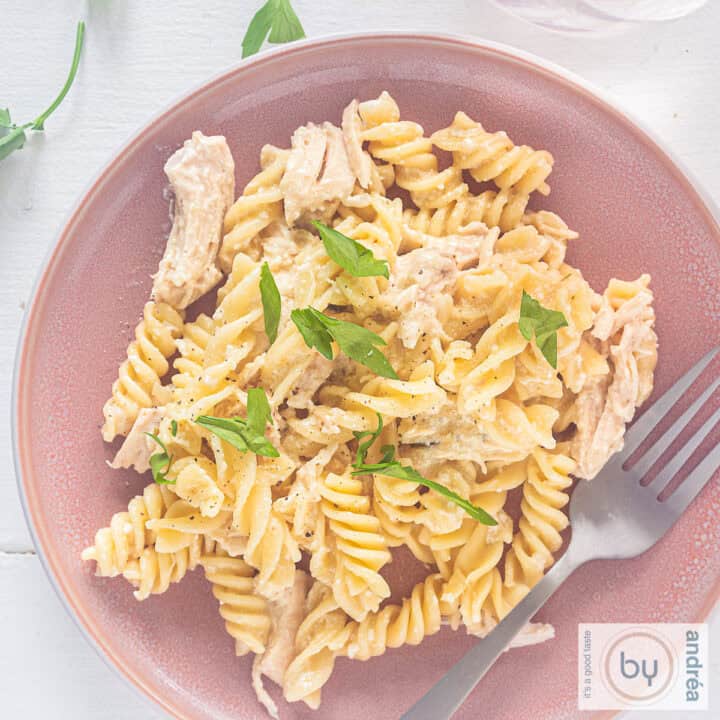 Een vierkante foto van een roze bord met pasta, romige saus met Parmezaan en knoflook en peterselie op een witte ondergrond