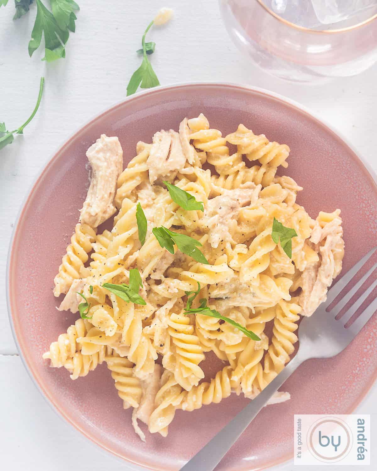 Een foto van bovenaf van een roze bord met pasta, kip en een romige Parmezaanse knoflooksaus. Een witte ondergrond.