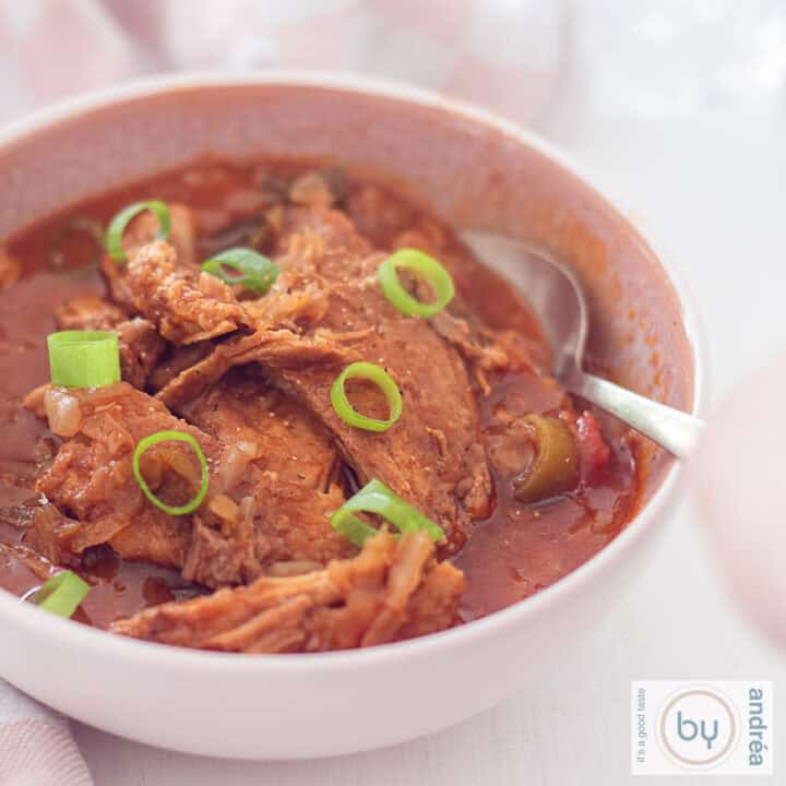 Een vierkante foto met een roze kom gevuld met hamlappen, groenten en tomatensaus. Bestrooid met lente ui. Een lepel ligt erin.