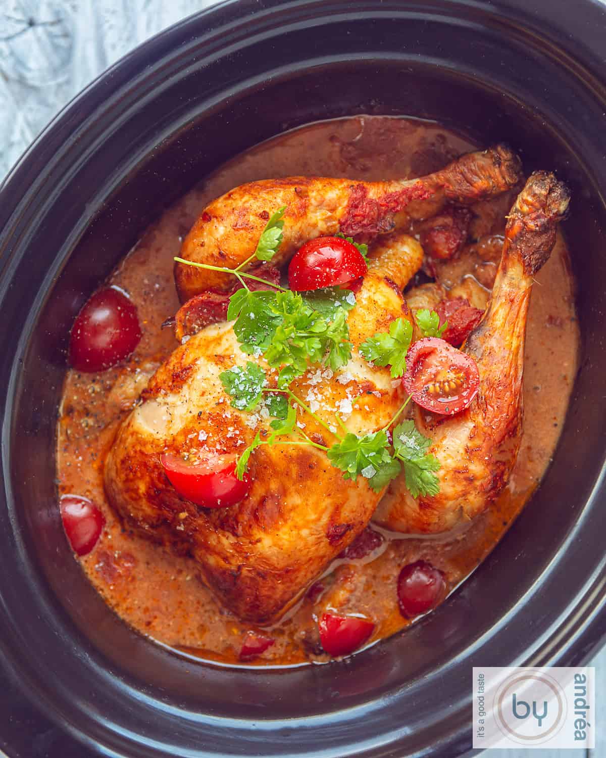 Een foto van bovenaf in een slowcooker met hele kip, romige saus, peterselie en cherry tomaten.