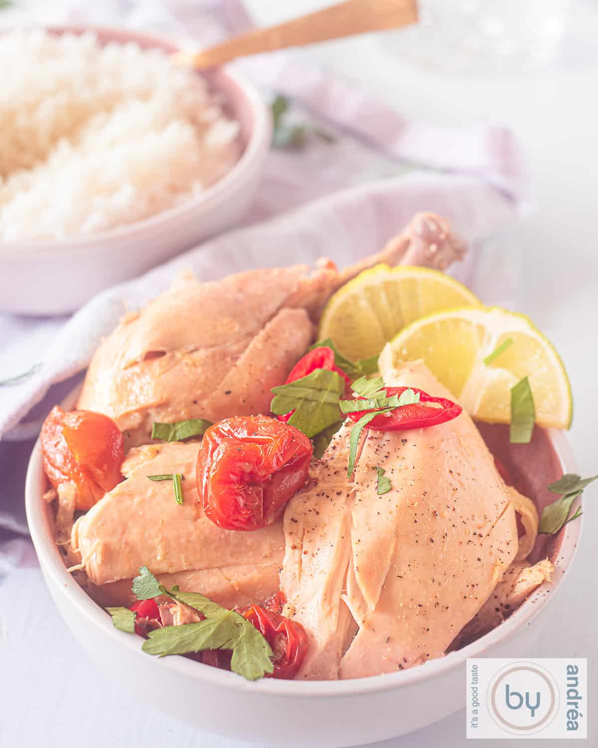 Een hoogtefoto met een roze schaal met stukken kip met romige curry saus met tomaat, citroen en peterselie. In de achtergrond een kommetje met witte rijst.