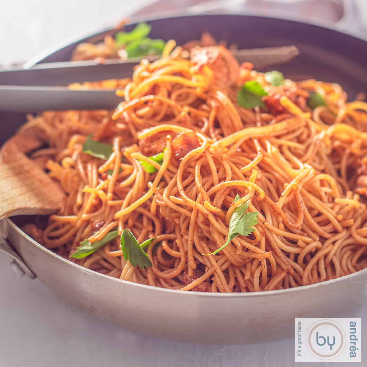 Een vierkante foto met een wok gevuld met Bami Surinaamse stijl met kip en snijselderij.