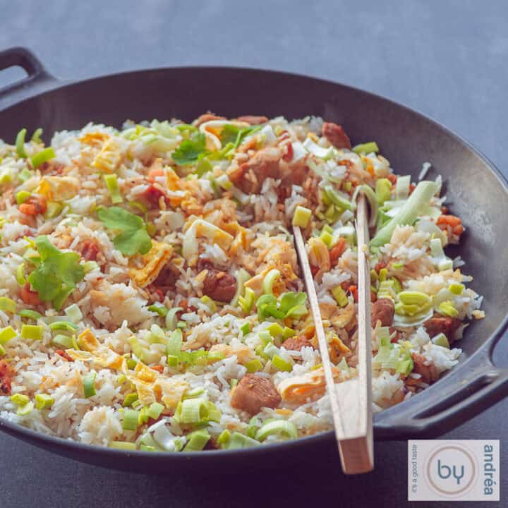 Een vierkante foto met een wok gevuld met nasi goreng met ham, kip en spekjes en groenten. Een houten knijptang ligt in de wok. Een donkere achtergrond
