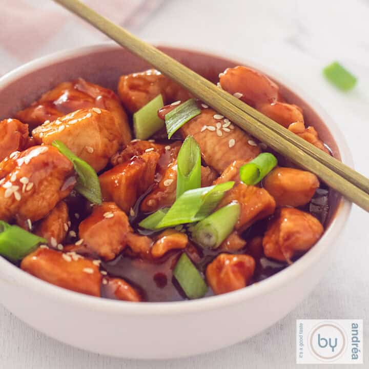 Een vierkante foto met een roze kom gevuld met kip, sticky saus, lente ui en sesam. Twee chopsticks liggen erbovenop.