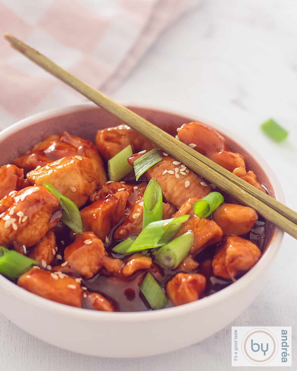 An aerial shot of a pink bowl of sticky chicken, spring onion and sesame seeds. Two golden chopsticks are placed on top.