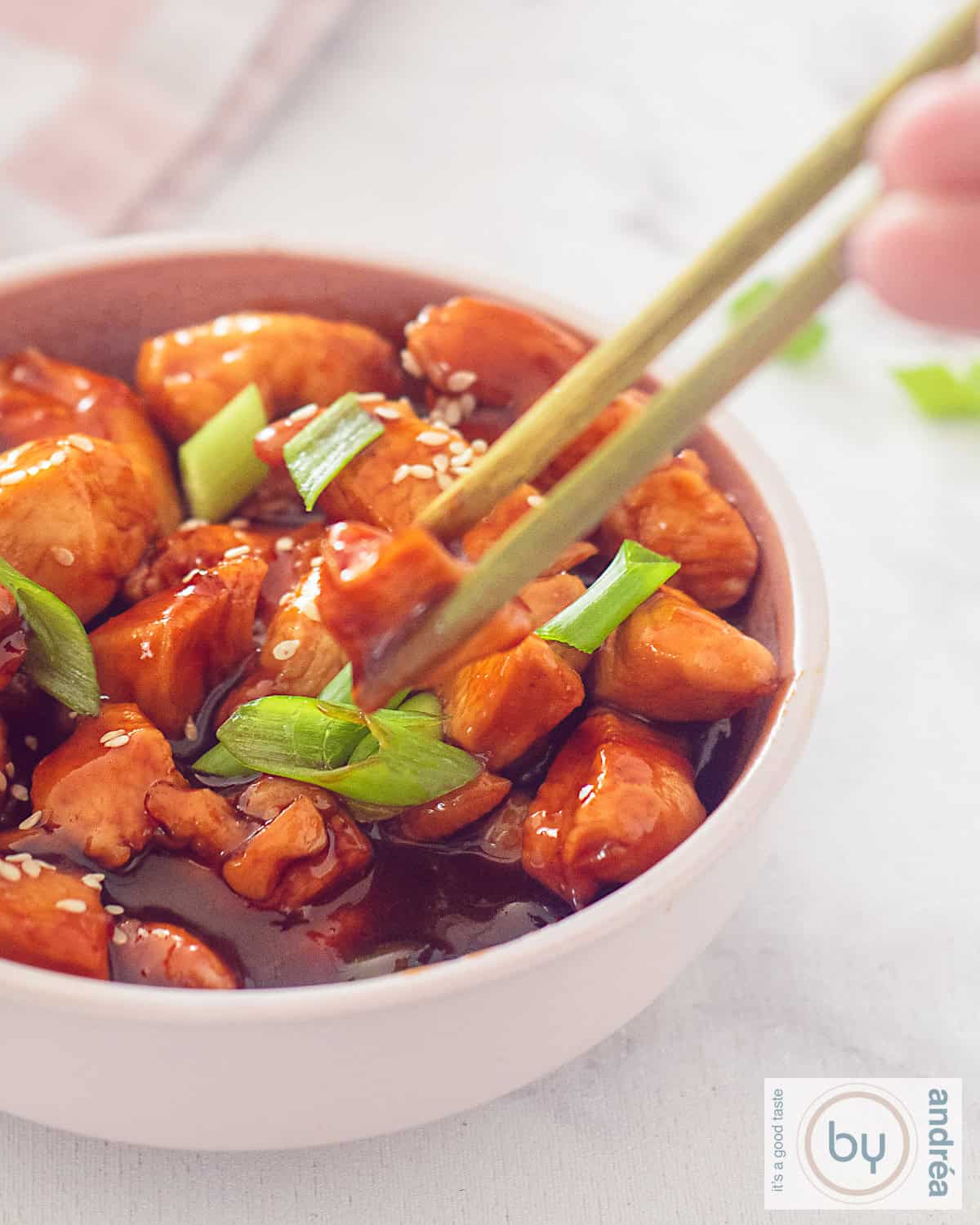 a portion of a pink bowl of sticky chicken with a garnish of spring onions and sesame. Two chopsticks grab a piece of chicken.