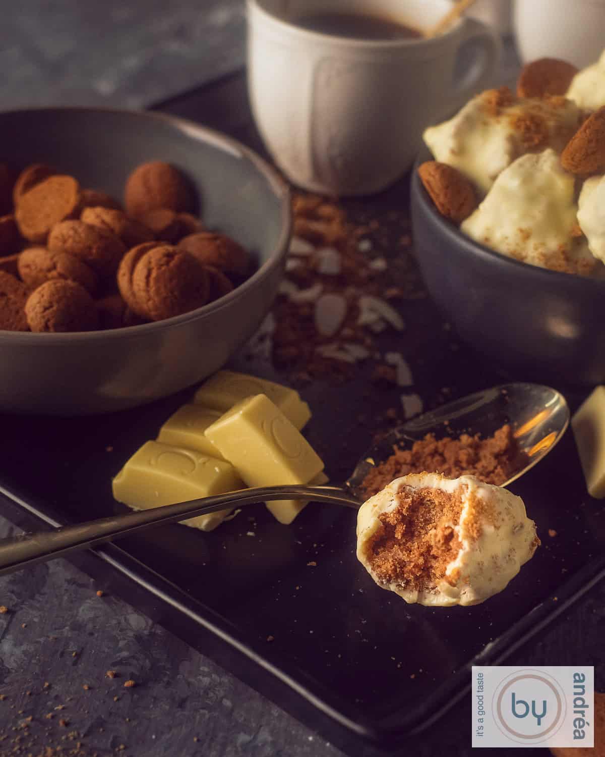 An height photo with a white chocolate truffle where the inside of cream cheese with pepernoten can be seen on a dark background. In the background, white chocolate, gingerbread and more truffles.