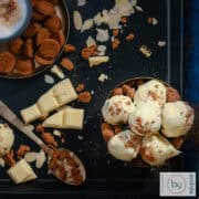 Een vierkante foto van een zwarte ondergrond met een kom gevuld met pepernoten truffels met witte chocola, daaromheen pepernoten, witte chocola, amandelschaafsel en koffie.