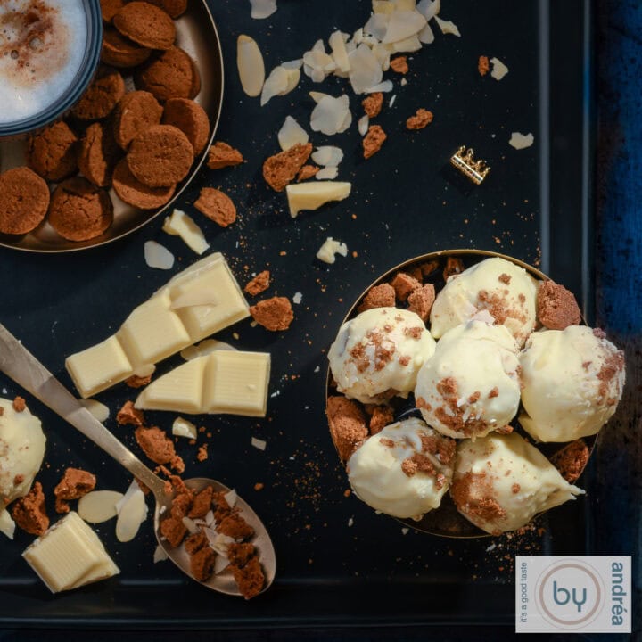 Een vierkante foto van een zwarte ondergrond met een kom gevuld met pepernoten truffels met witte chocola, daaromheen pepernoten, witte chocola, amandelschaafsel en koffie.