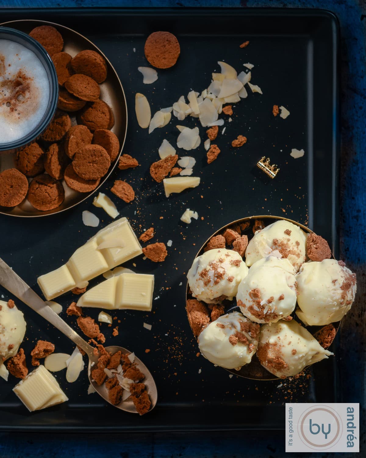 A photo from above of a black background with a bowl of white chocolate pepernoten truffles, a bowl of gingerbread, white chocolate, and almond shavings