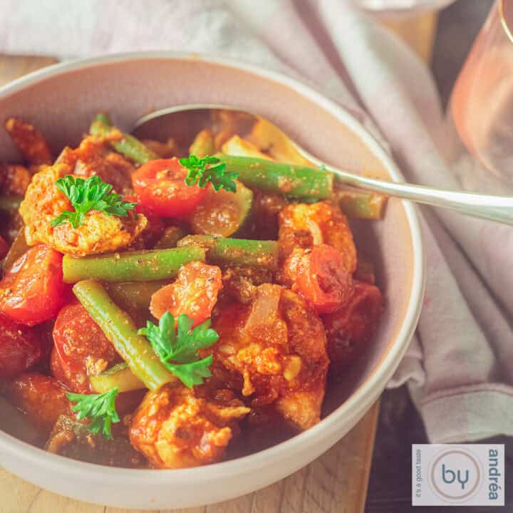 Een vierkante foto met een roze schaal met kipblokjes, boontjes, tomaat en courgette in een kruidige saus.