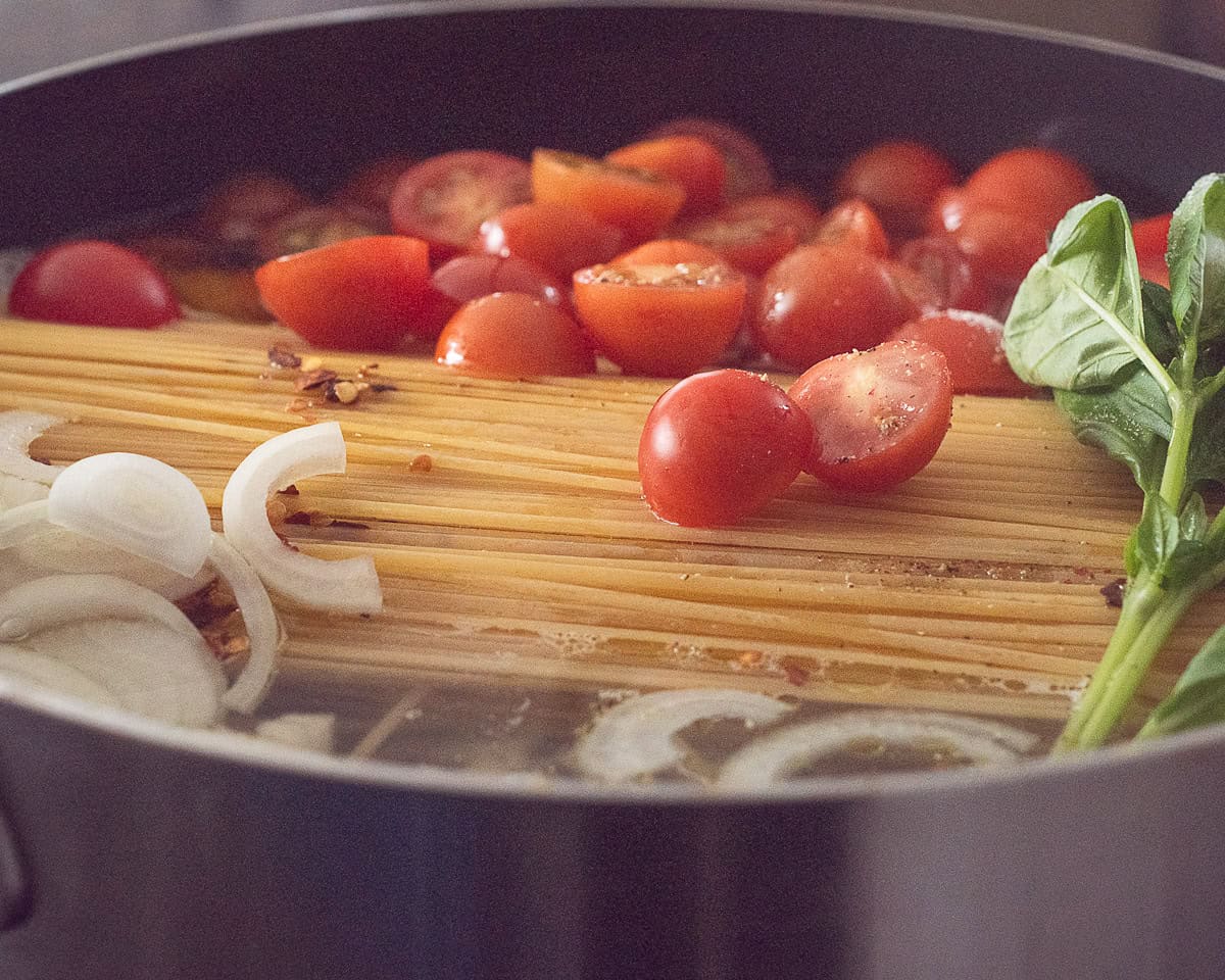 Een hapjespan gevuld met pasta, ui, tomaten, basilicum en water.