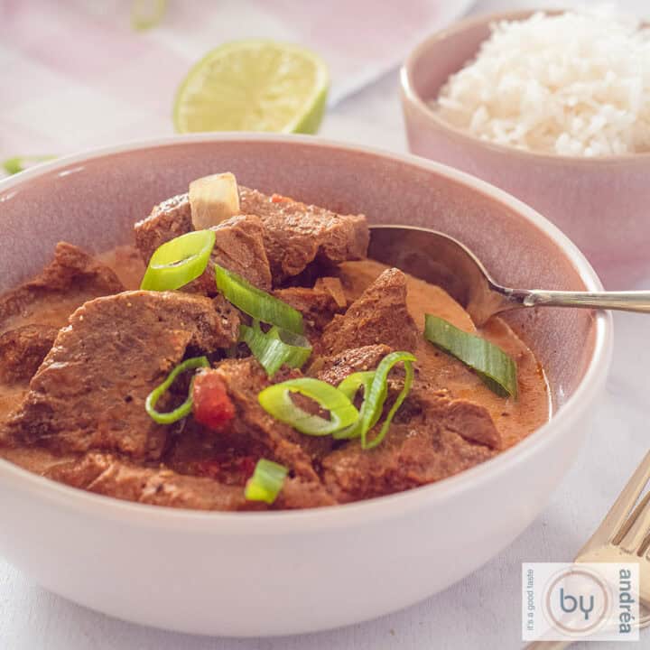 Een vierkante kom gevuld met curry madras en lente ui. Een roze kom gevuld met rijst er schuin achter.