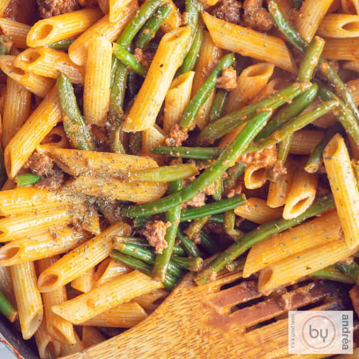 Een vierkante foto ment een pan gevuld met penne alla vodka, met sperziebonen en gehakt. Een houten lepel ligt in de pan.
