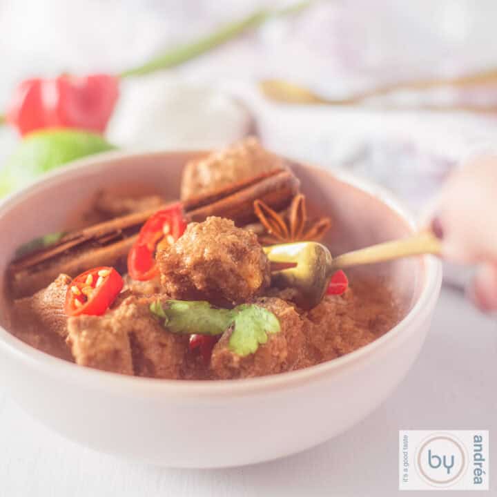 Een vierkante foto met een roze kom gevuld met rendang beef, kaneelstokje, peterselie en rode peper. Een hand houdt een vork vast.