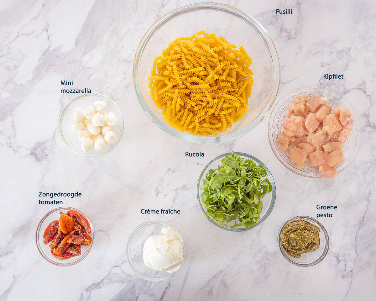 Ingredienten Pasta Pesto met Kip in 15 Minuten op een wit marmeren ondergrond met tekstbeschrijving.
