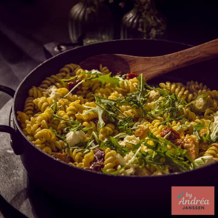Een vierkante foto met een zwarte pan met pasta pesto met kip, rucola en zongedroogde tomaten.
