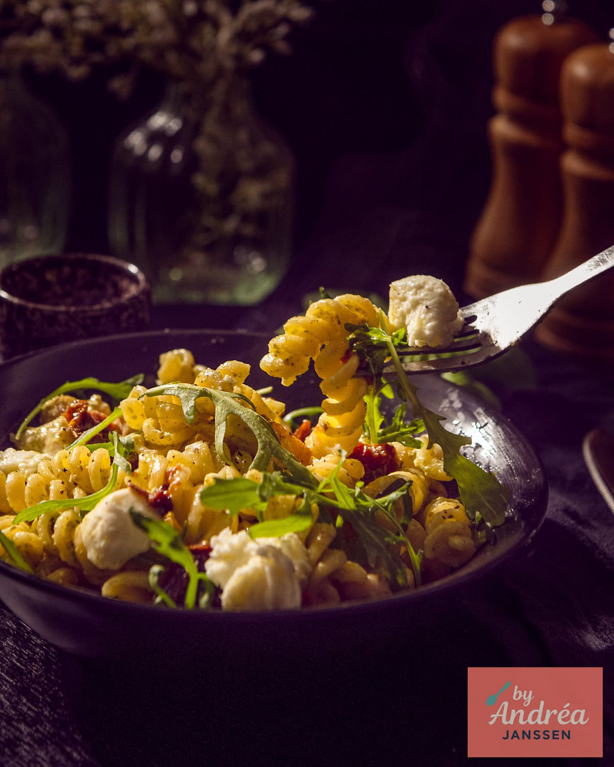 Een schaaltje met pasta met pestosaus, rucola, mozzarella, zongedroogde tomaten en kip. Een vork neemt een hap