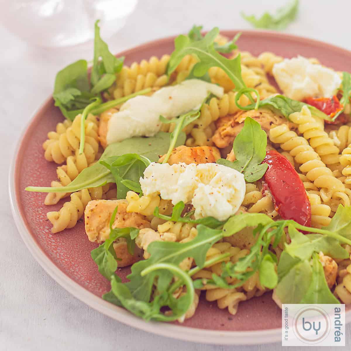 Een vierkante foto met een roze bord met fusilli met zongedroogde tomaten, rucola, mozzarella en pestosaus