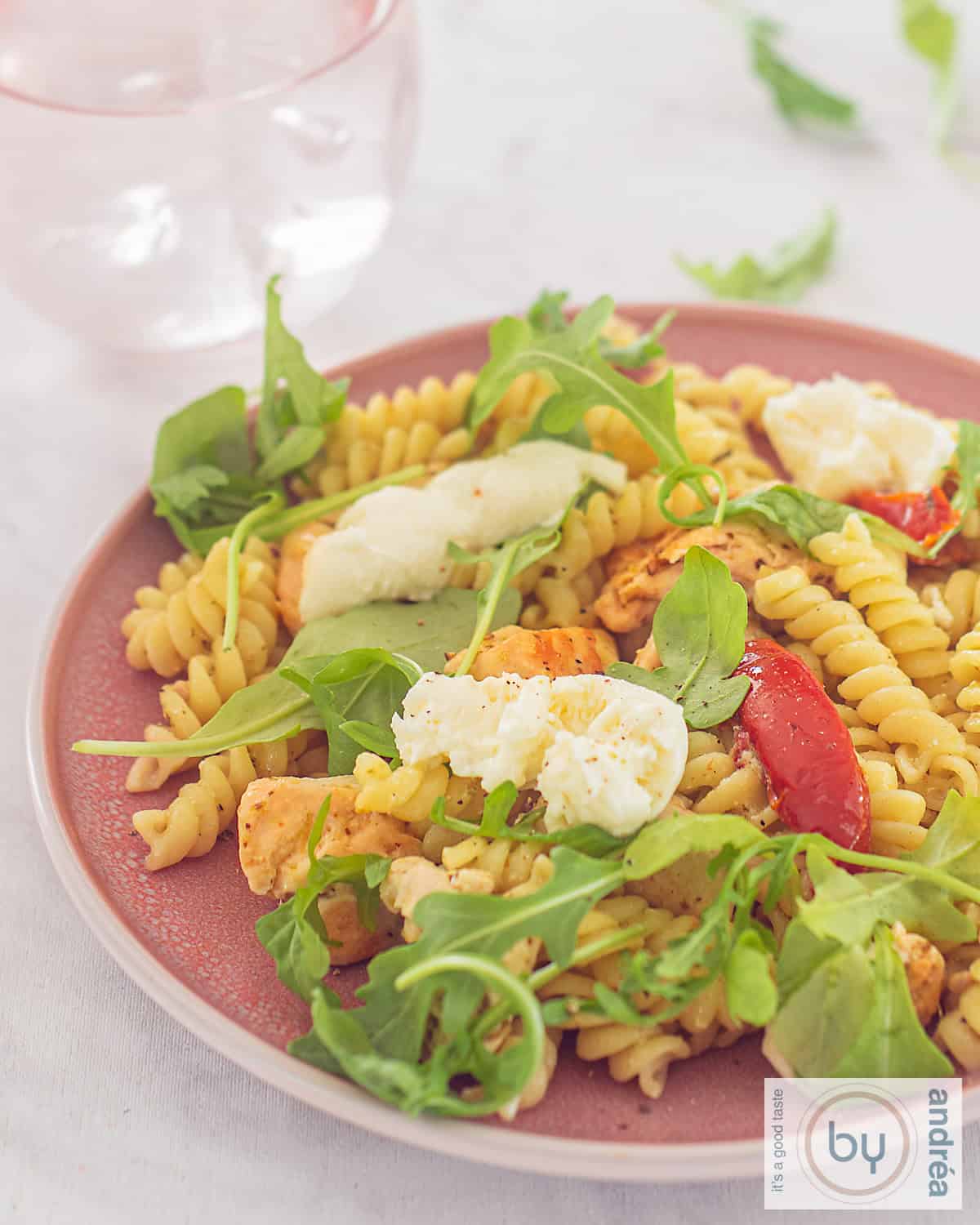 Een foto van een deel van een roze bord met fusilli, pesto, mozzarella en rucola met zongedroogde tomaten.