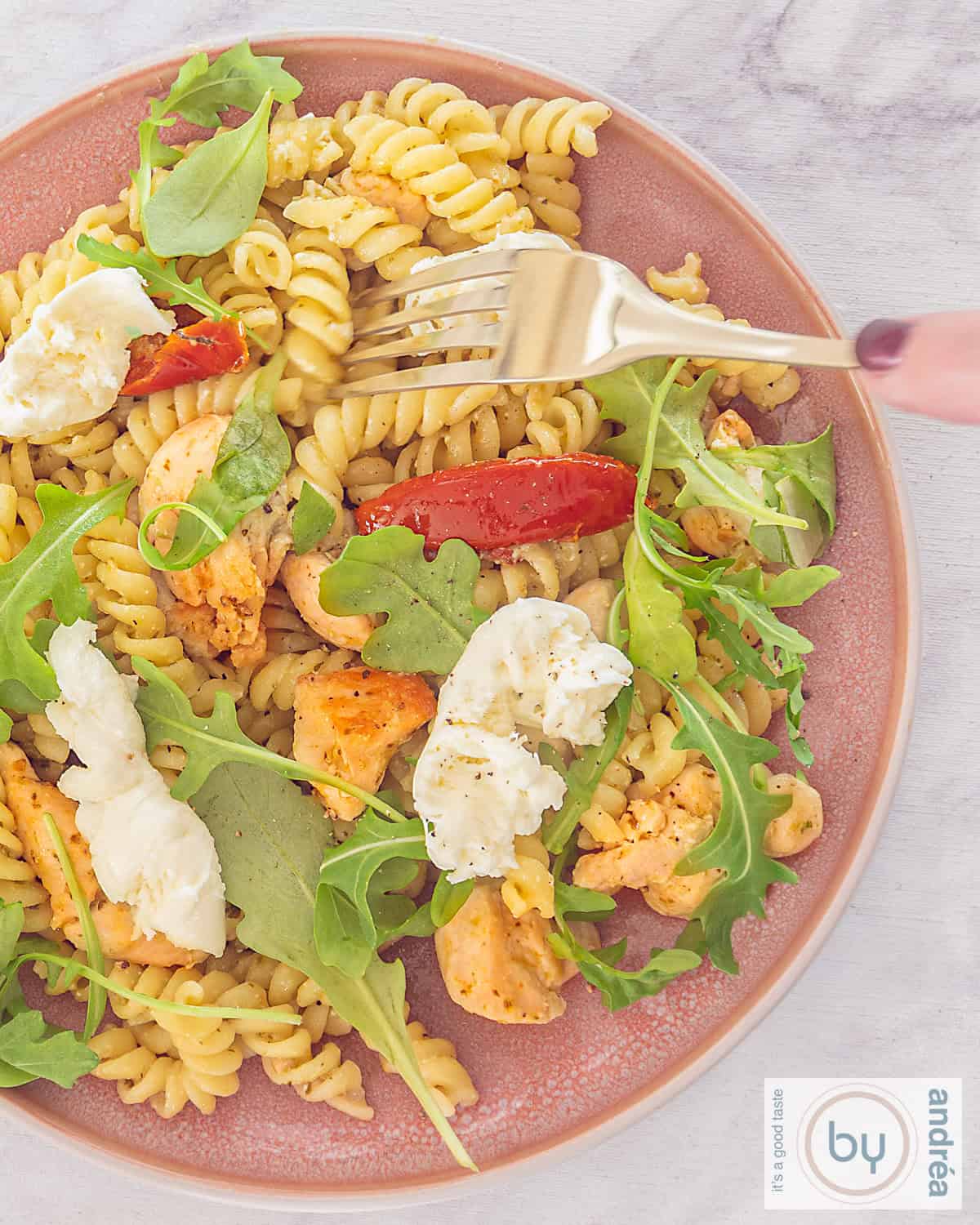 Een foto van bovenaf van een roze bord met pasta pesto met kip, mozzarella en zongedroogde tomaten met rucola.