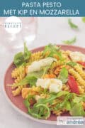 Een roze bord met pesto pasta met kip, mozzarella, rucola en zongedroogde tomaten. Een tekst ovenin: pasta pesto met kip en mozzarella