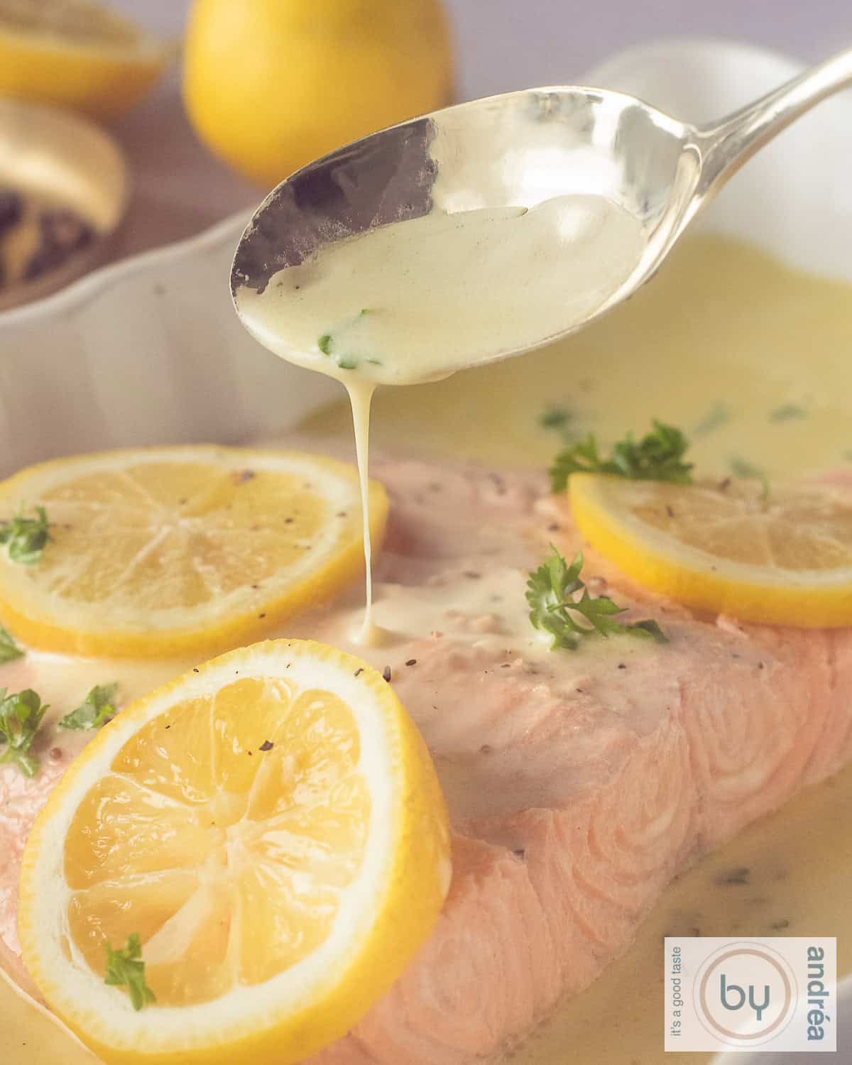 Een hoogtefoto gepocheerde zalm in witte wijnsaus. Een lepel schenkt saus op de vis. Er liggen plakken citroen en peterselie op de vis.