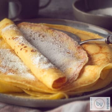 Een vierkante foto met een donkergrijs bord met een stapel versgebakken pannenkoeken met poedersuiker.