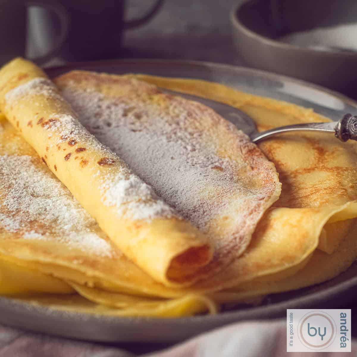 Een vierkante foto met een donkergrijs bord met een stapel versgebakken pannenkoeken met poedersuiker.