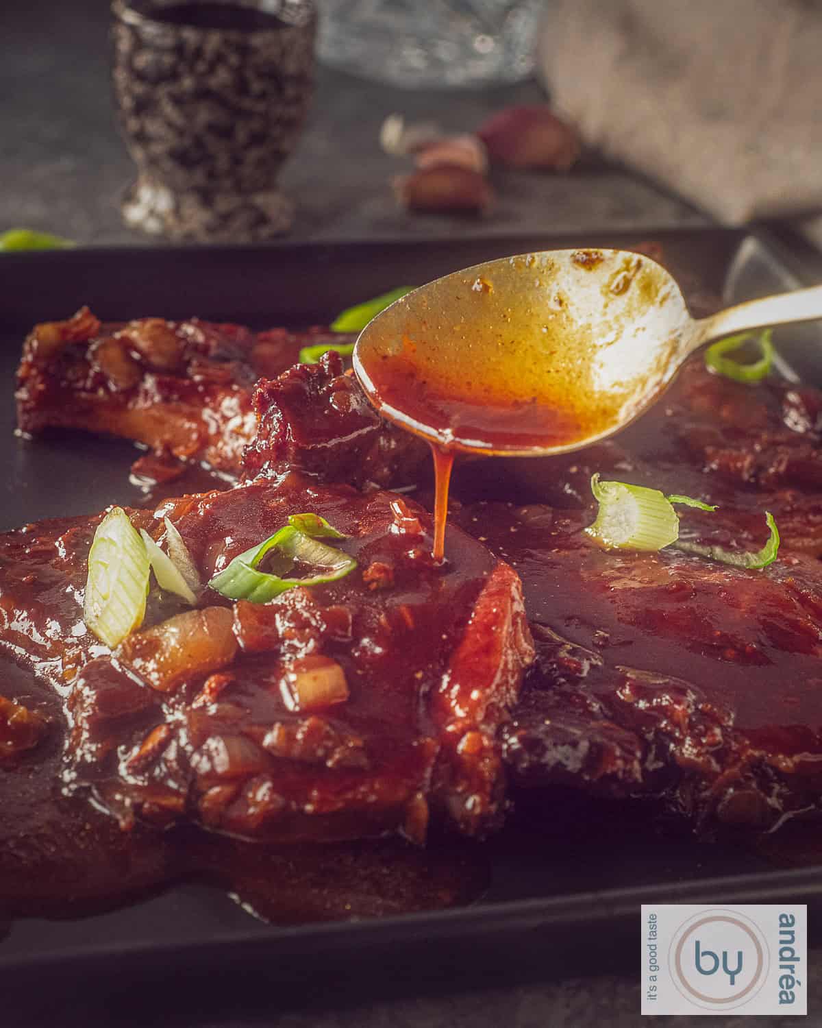 Een vierkant zwart bord emt karbonades met saus. Een lepel schenkt sojasaus op het vlees.